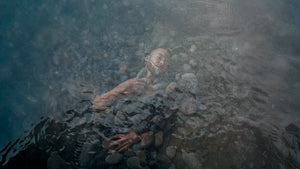 Astrologer Jeramie Bellmay submerged under calm rippling water, partially buried beneath ocean tumbled stones